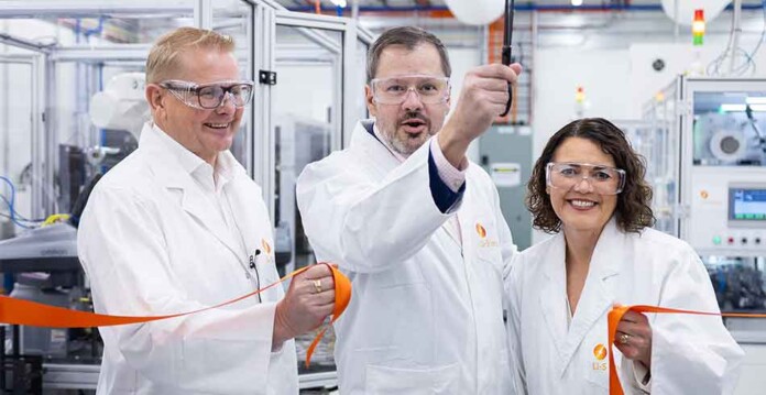 Two men and a woman wearing lab coats cut ribbon at opening of battery manufacturing facility