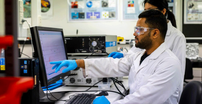 Male and female scientists in lab coats analyse information on computer screen (li-s energy)